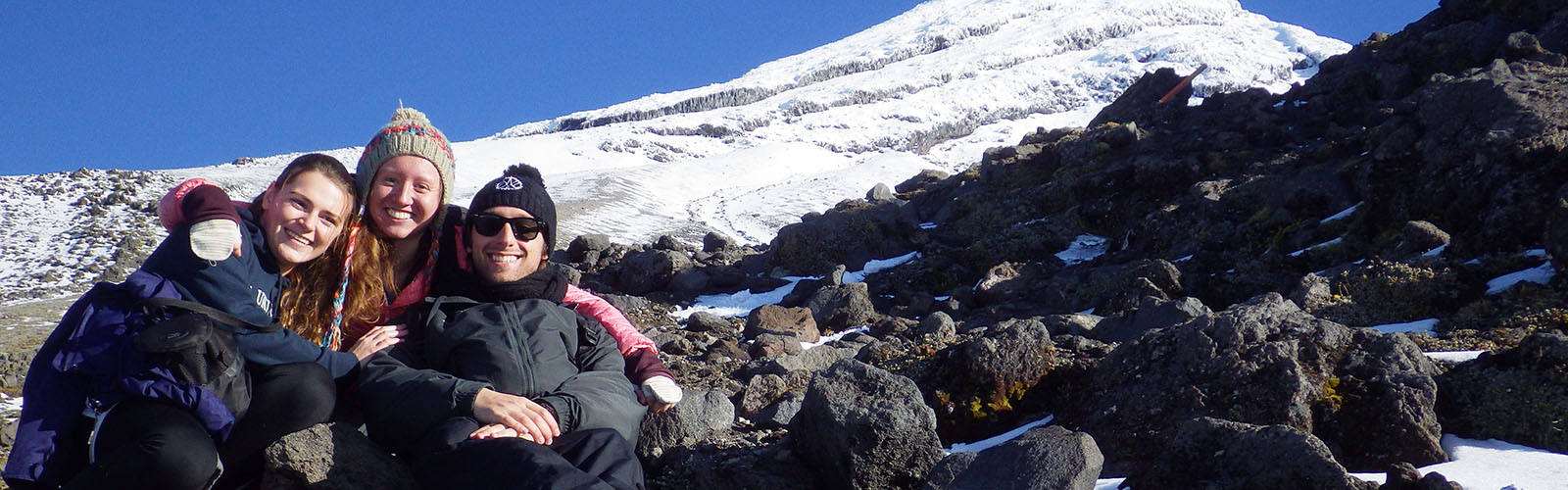 Three students in warm clothing are standing on a rocky edge of a snow-covered mountain.